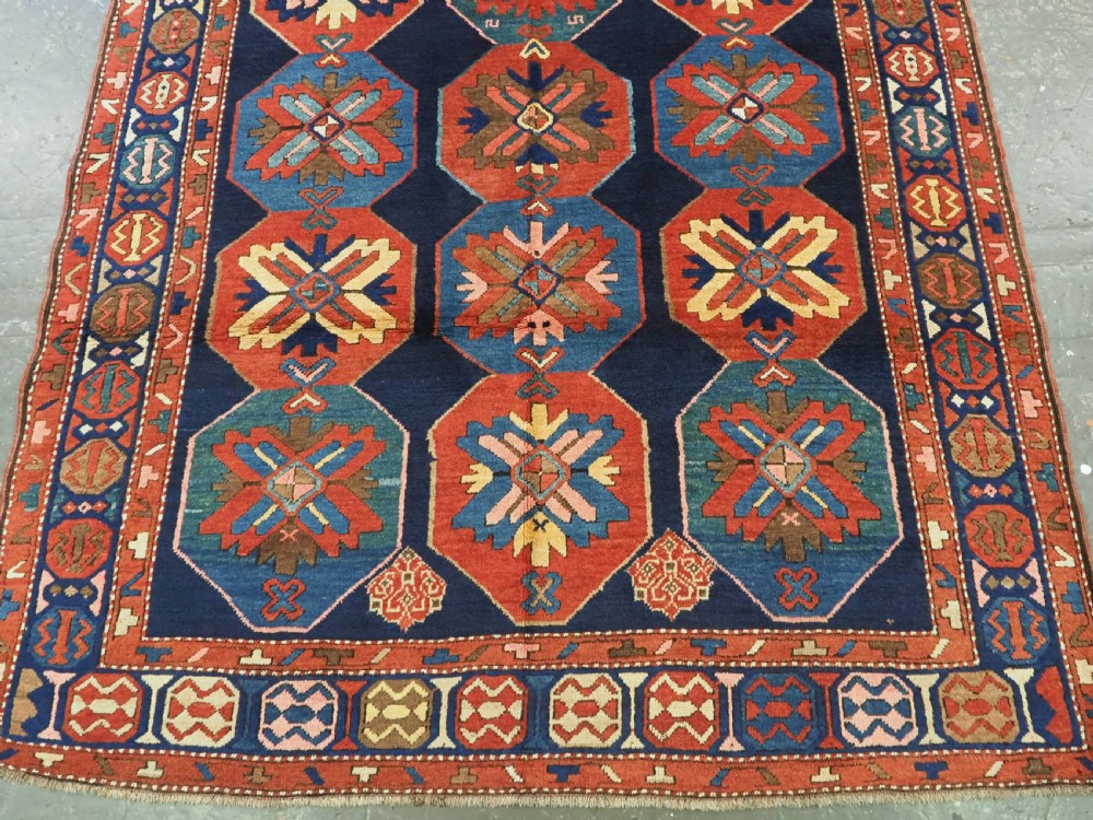 An antique hand-knotted wool rug featuring columns of red and blue hexagonal medallions on a deep navy field framed by a geometric multi-band border. View VII