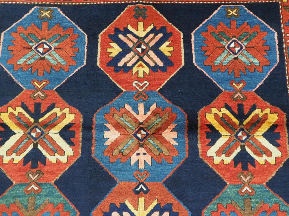 An antique hand-knotted wool rug featuring columns of red and blue hexagonal medallions on a deep navy field framed by a geometric multi-band border. View VIII