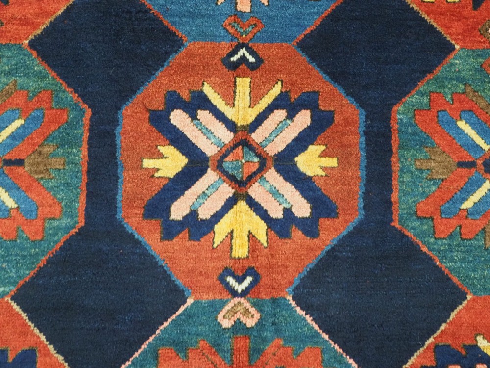 An antique hand-knotted wool rug featuring columns of red and blue hexagonal medallions on a deep navy field framed by a geometric multi-band border. View IX