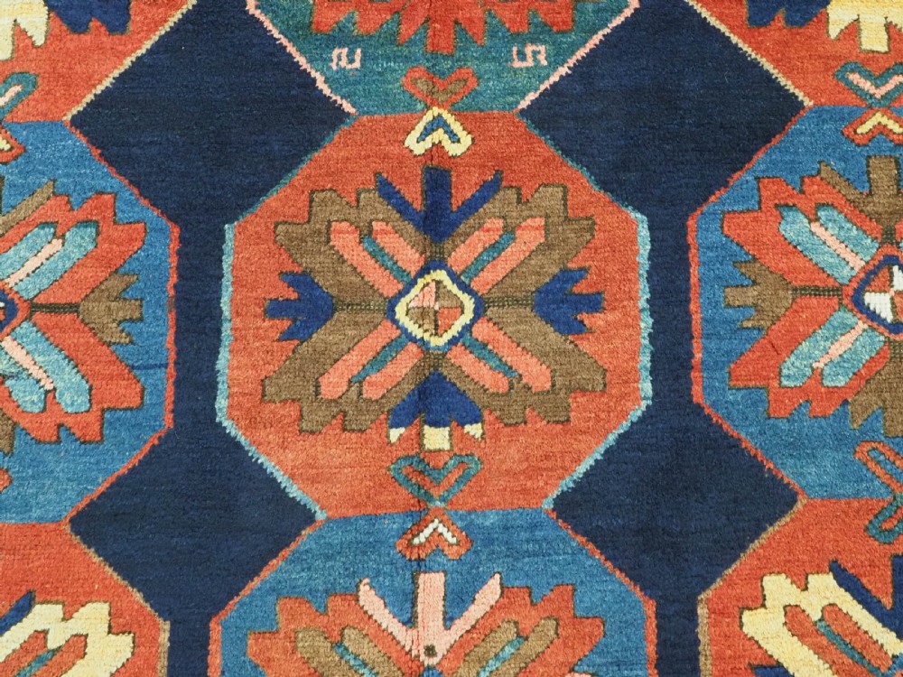 An antique hand-knotted wool rug featuring columns of red and blue hexagonal medallions on a deep navy field framed by a geometric multi-band border. View X
