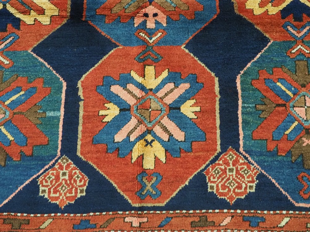 An antique hand-knotted wool rug featuring columns of red and blue hexagonal medallions on a deep navy field framed by a geometric multi-band border. View XI