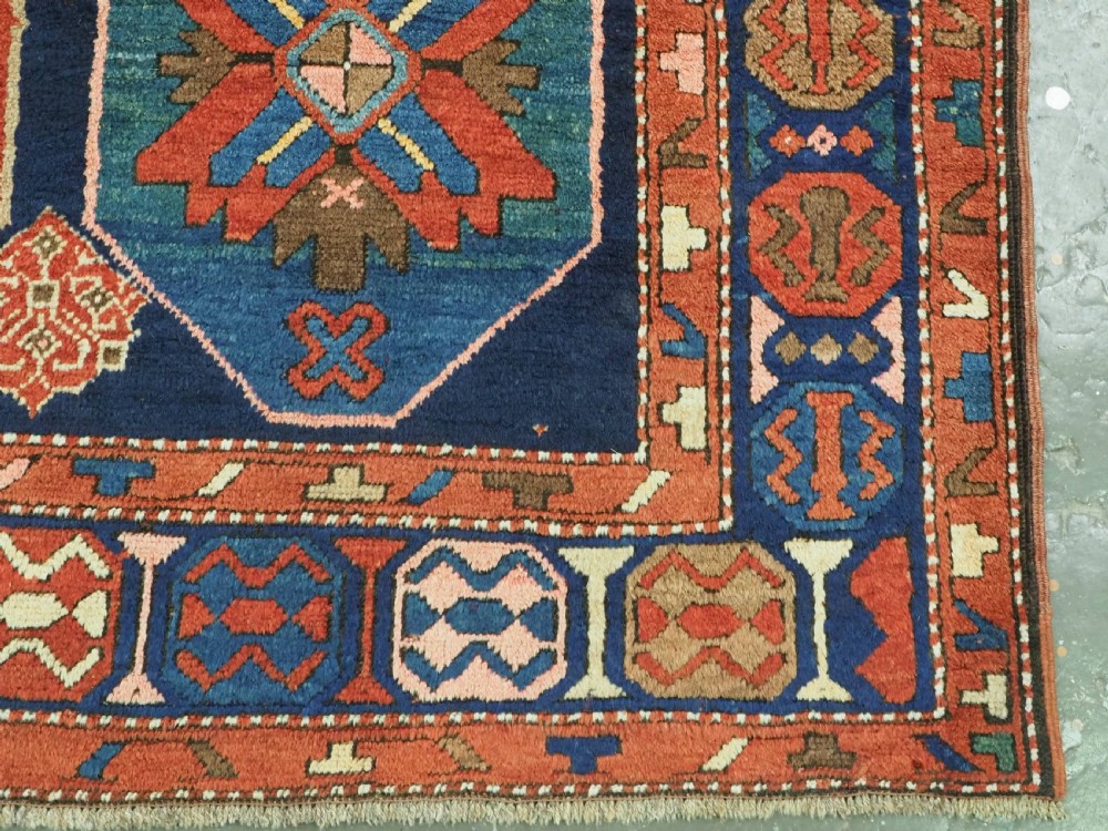 An antique hand-knotted wool rug featuring columns of red and blue hexagonal medallions on a deep navy field framed by a geometric multi-band border. View XII