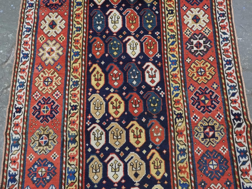 An antique hand-knotted Caucasian runner rug featuring repeated boteh motifs on a dark blue central field framed by red geometric borders. View III