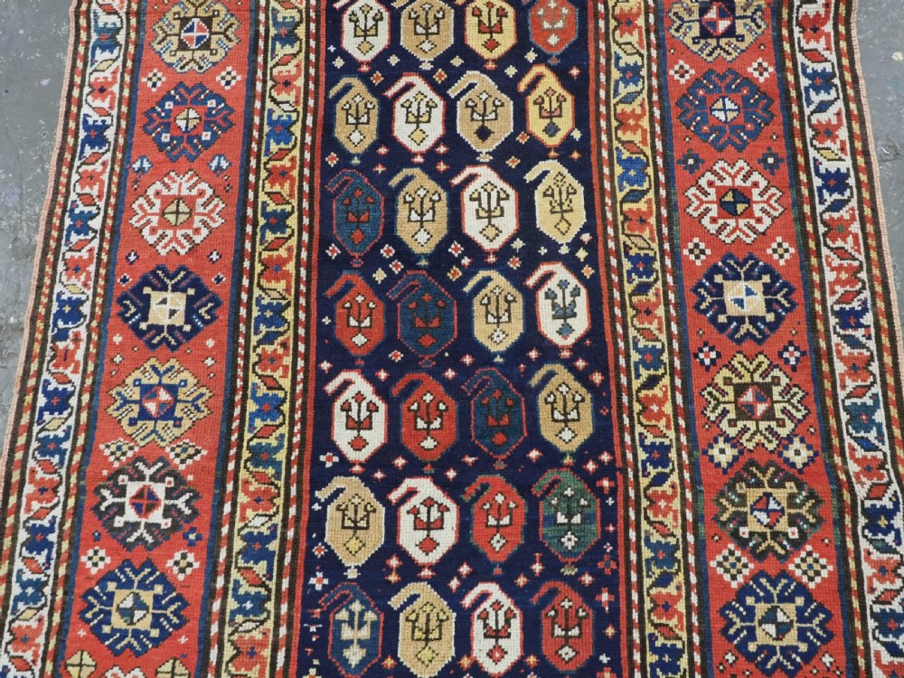 An antique hand-knotted Caucasian runner rug featuring repeated boteh motifs on a dark blue central field framed by red geometric borders. View IV