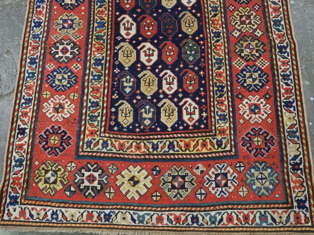 An antique hand-knotted Caucasian runner rug featuring repeated boteh motifs on a dark blue central field framed by red geometric borders. View V