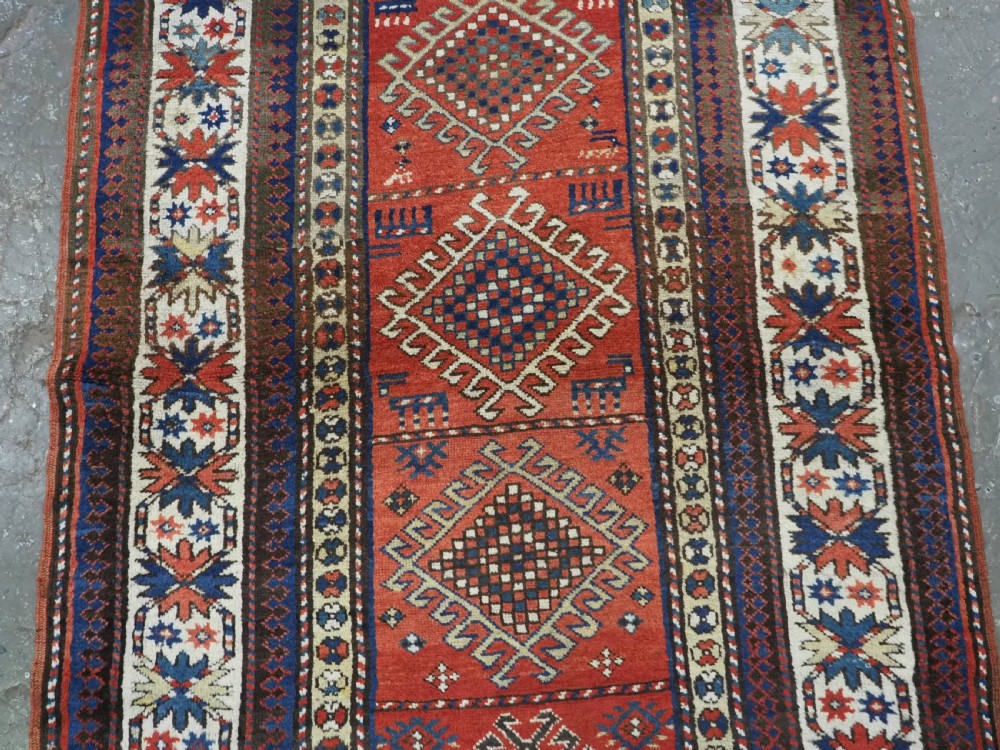 An antique hand-knotted Caucasian Moghan Kazak wool runner featuring four central hooked diamond medallions on a red field enclosed by multi-band geometric borders. View V