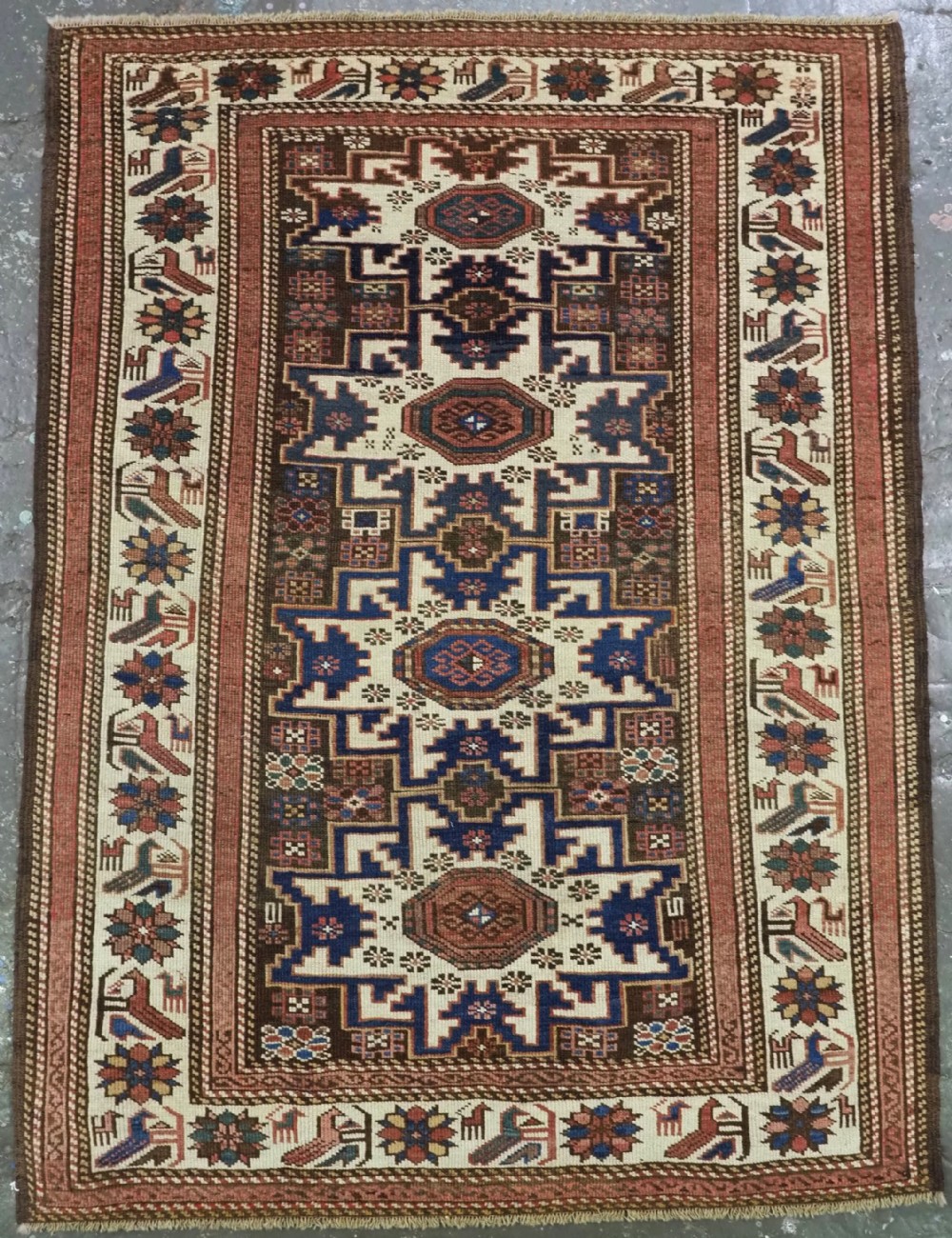 A hand-knotted antique Caucasian wool rug featuring geometric Lesghi star medallions on a dark central field enclosed by an ivory border decorated with stylised bird motifs.