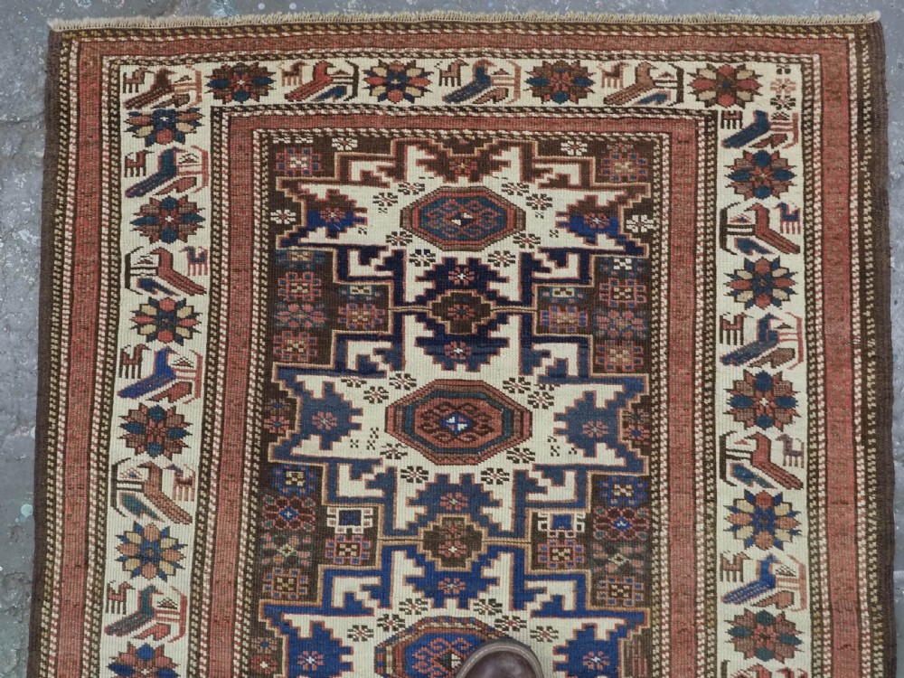 A hand-knotted antique Caucasian wool rug featuring geometric Lesghi star medallions on a dark central field enclosed by an ivory border decorated with stylised bird motifs. View III
