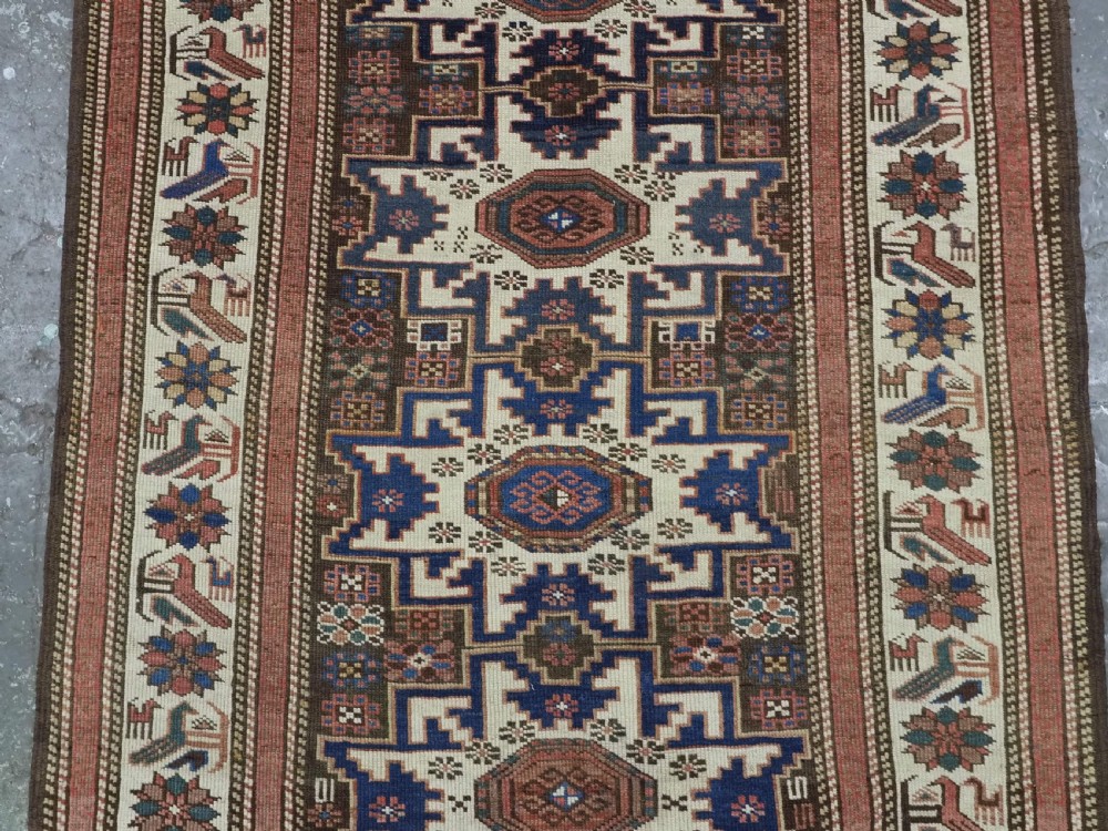 A hand-knotted antique Caucasian wool rug featuring geometric Lesghi star medallions on a dark central field enclosed by an ivory border decorated with stylised bird motifs. View IV