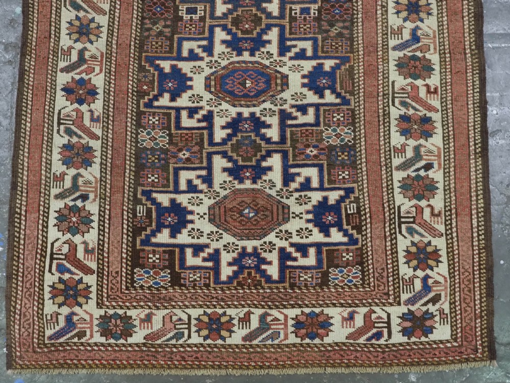 A hand-knotted antique Caucasian wool rug featuring geometric Lesghi star medallions on a dark central field enclosed by an ivory border decorated with stylised bird motifs. View V