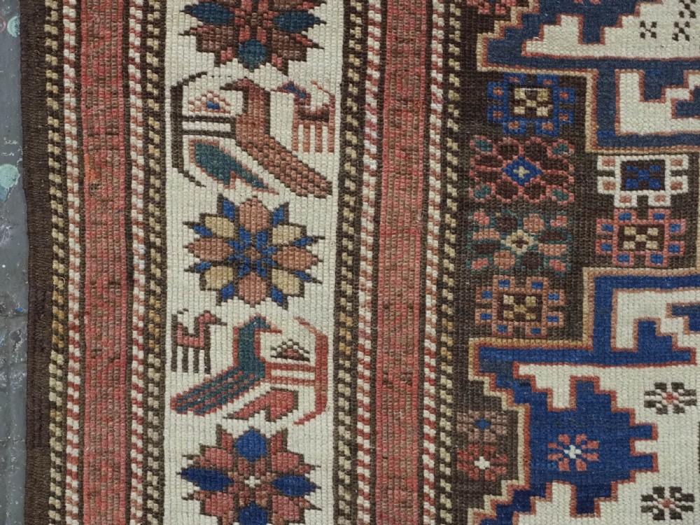 A hand-knotted antique Caucasian wool rug featuring geometric Lesghi star medallions on a dark central field enclosed by an ivory border decorated with stylised bird motifs. View VII