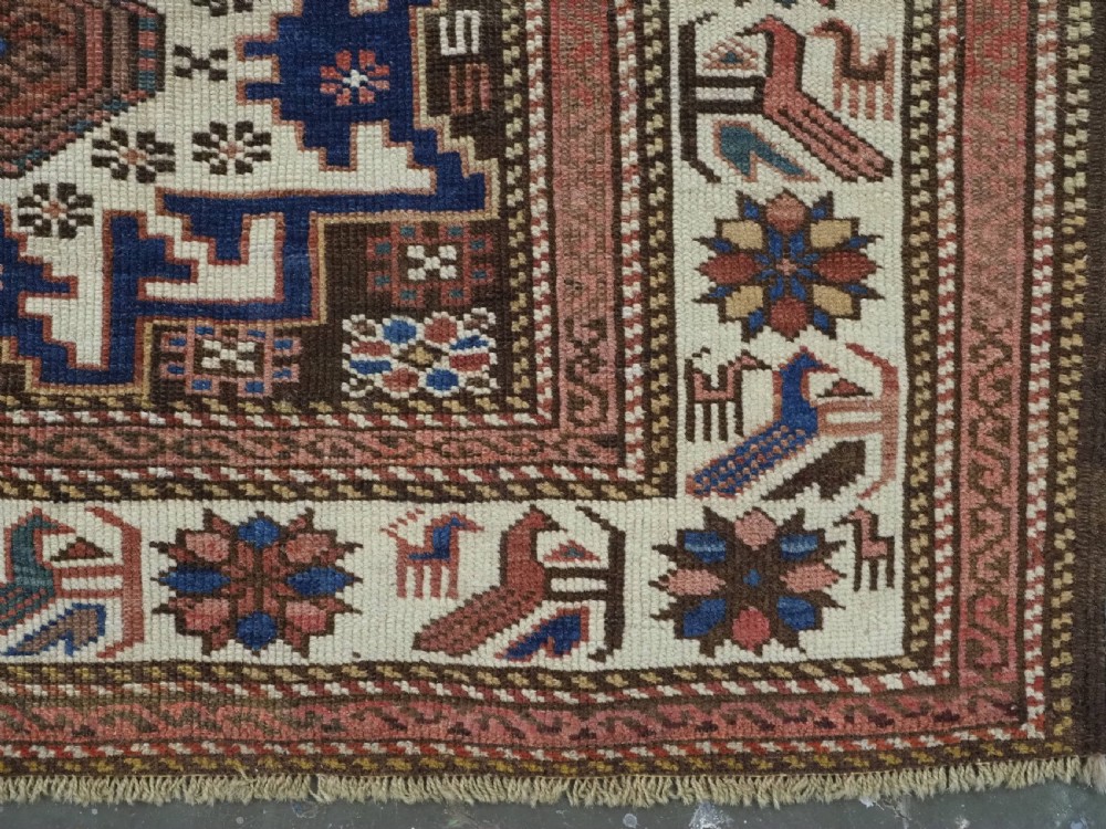 A hand-knotted antique Caucasian wool rug featuring geometric Lesghi star medallions on a dark central field enclosed by an ivory border decorated with stylised bird motifs. View X