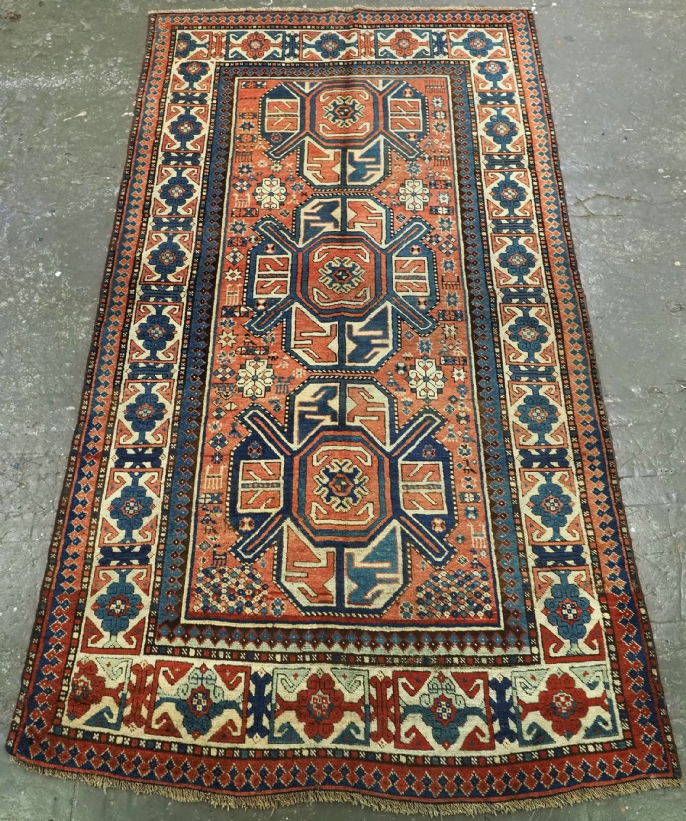 An antique hand-knotted Caucasian wool rug featuring three primary geometric medallions on a terracotta field with navy and cream patterned borders.