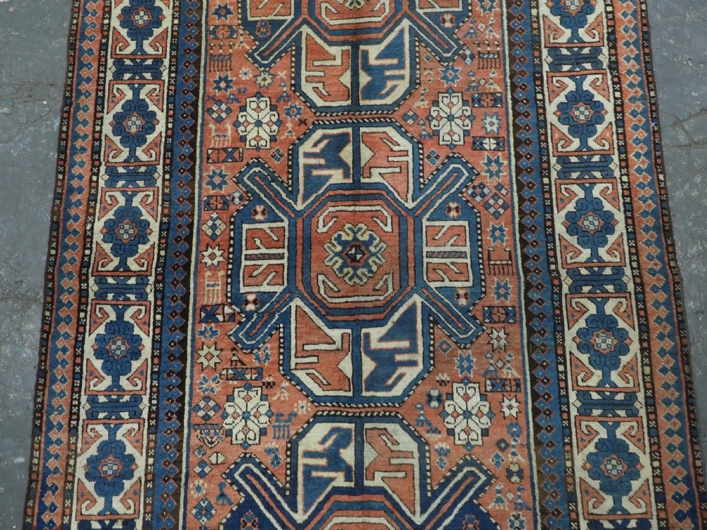 An antique hand-knotted Caucasian wool rug featuring three primary geometric medallions on a terracotta field with navy and cream patterned borders. View IV
