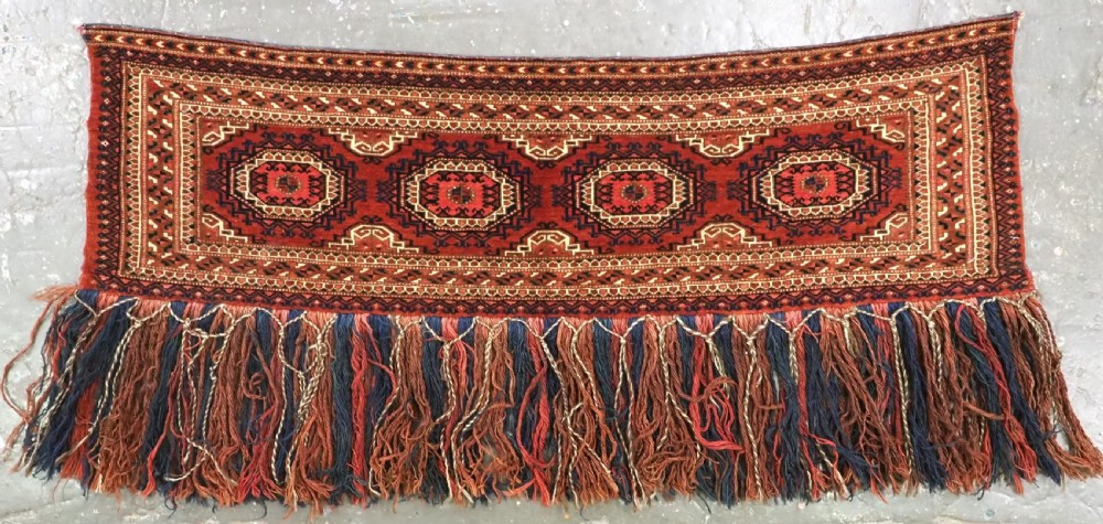 An antique rectangular handwoven Tekke Turkmen wool torba featuring four geometric octagonal guls on a deep red field with long multi-coloured fringe along the bottom edge.
