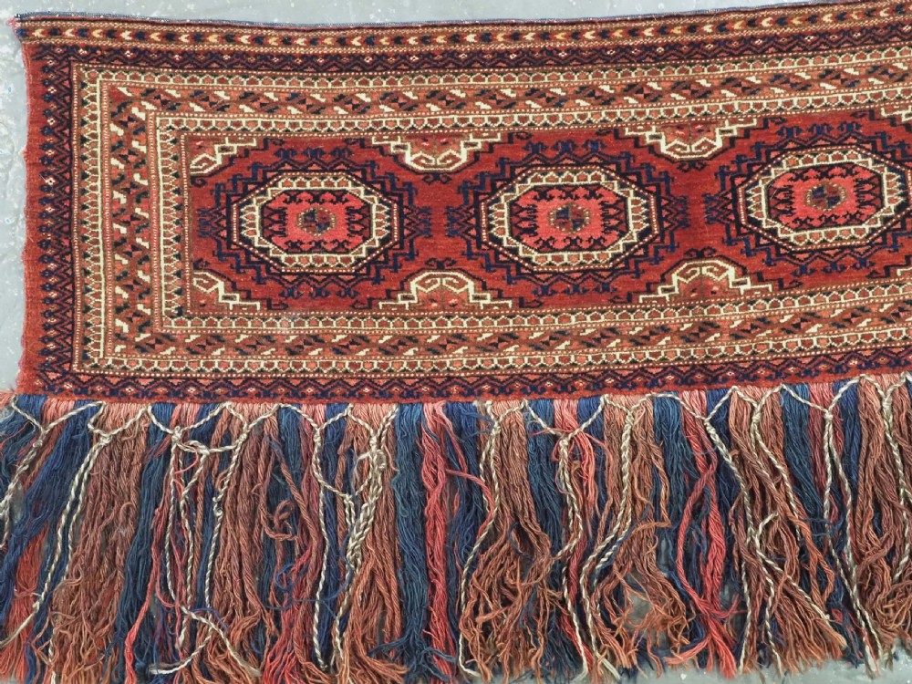 An antique rectangular handwoven Tekke Turkmen wool torba featuring four geometric octagonal guls on a deep red field with long multi-coloured fringe along the bottom edge. View II