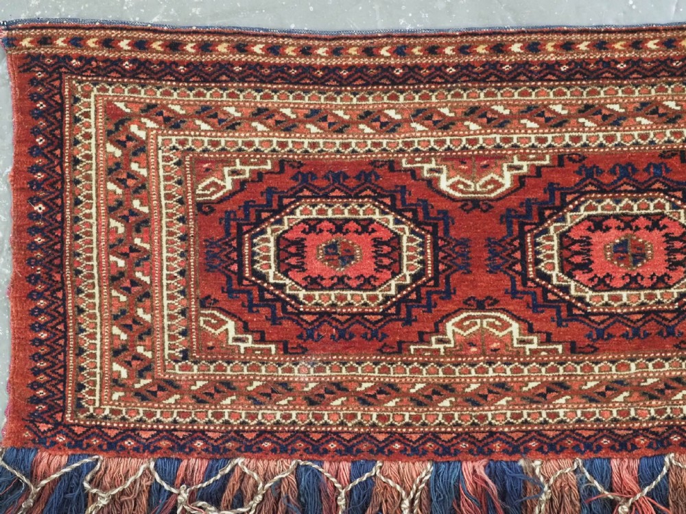 An antique rectangular handwoven Tekke Turkmen wool torba featuring four geometric octagonal guls on a deep red field with long multi-coloured fringe along the bottom edge. View III