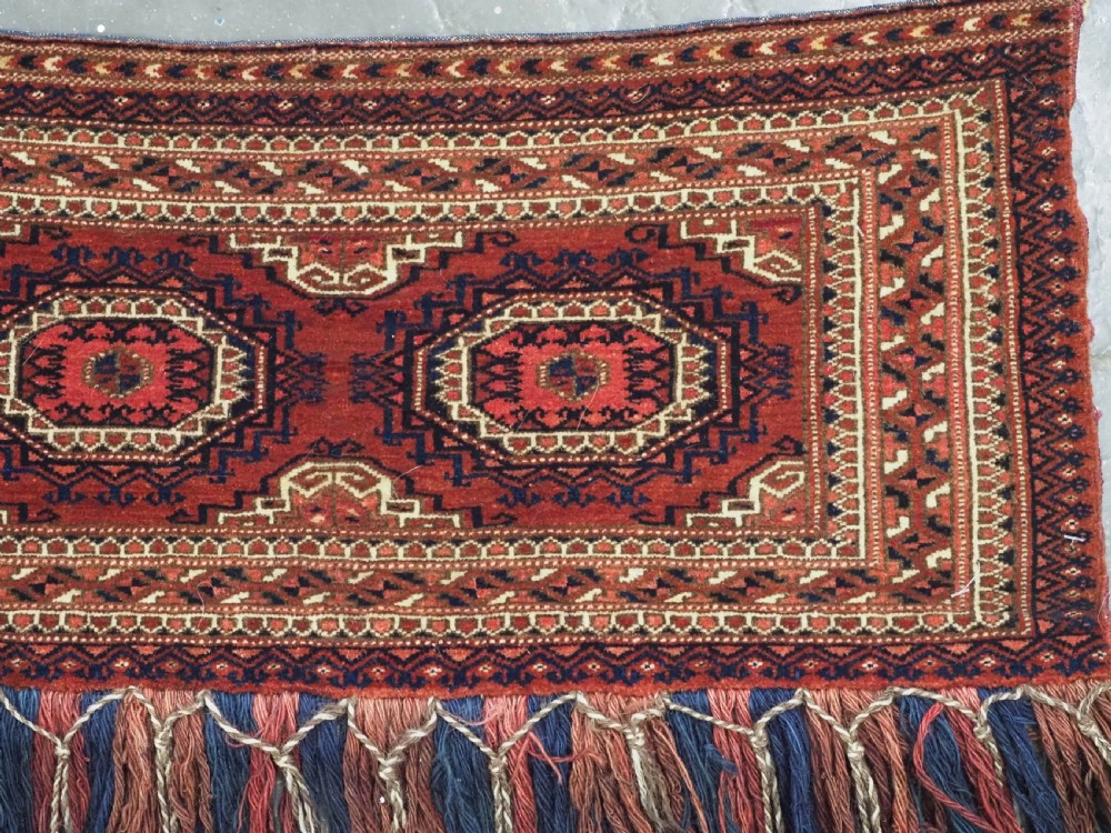 An antique rectangular handwoven Tekke Turkmen wool torba featuring four geometric octagonal guls on a deep red field with long multi-coloured fringe along the bottom edge. View IV
