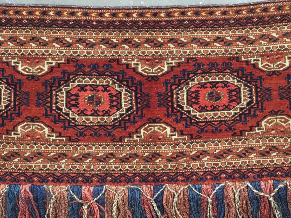 An antique rectangular handwoven Tekke Turkmen wool torba featuring four geometric octagonal guls on a deep red field with long multi-coloured fringe along the bottom edge. View V