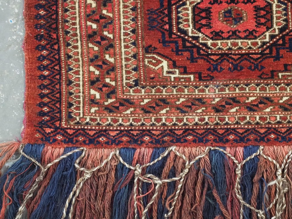 An antique rectangular handwoven Tekke Turkmen wool torba featuring four geometric octagonal guls on a deep red field with long multi-coloured fringe along the bottom edge. View VII