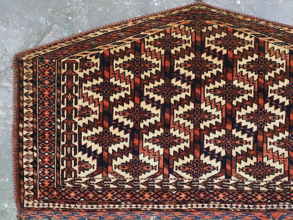 A pentagonal hand-knotted antique Yomut Turkmen Asmalyk wool textile featuring geometric lattice motifs in red, dark brown, and cream with hanging multi-coloured tassels along the lower edge. View III