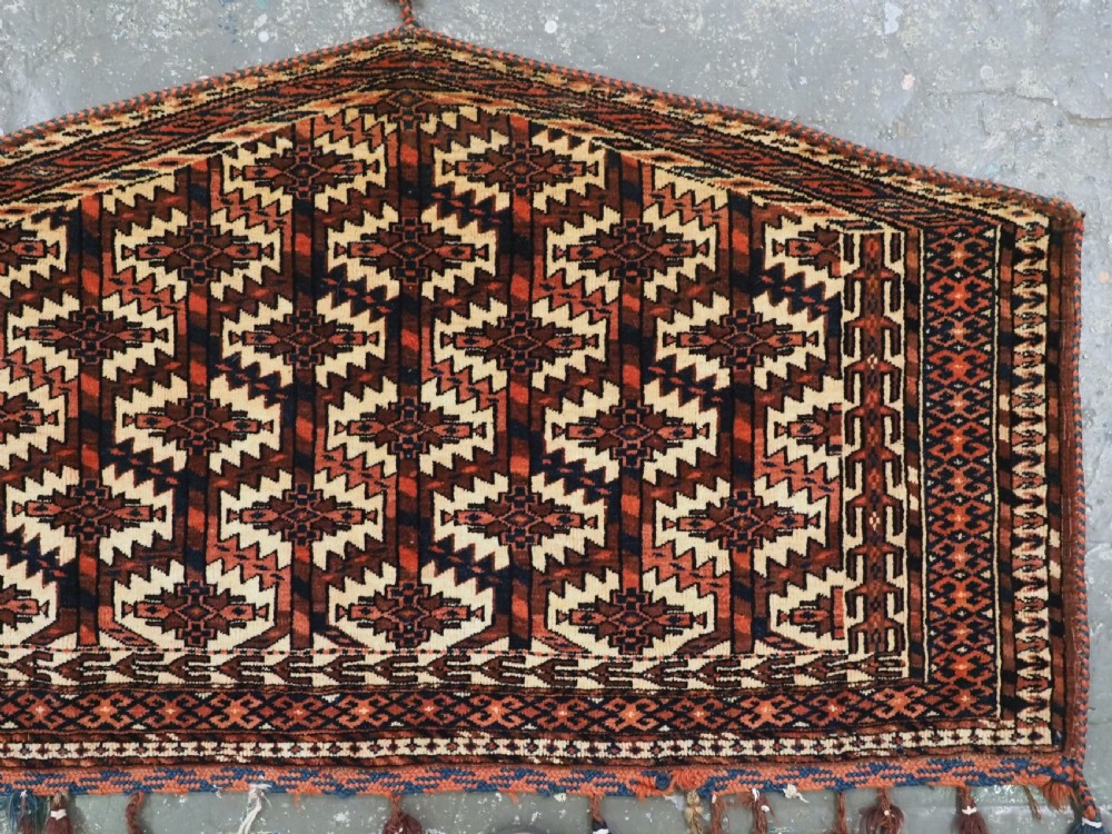 A pentagonal hand-knotted antique Yomut Turkmen Asmalyk wool textile featuring geometric lattice motifs in red, dark brown, and cream with hanging multi-coloured tassels along the lower edge. View IV