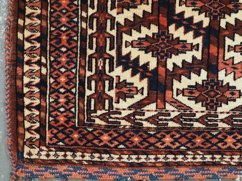 A pentagonal hand-knotted antique Yomut Turkmen Asmalyk wool textile featuring geometric lattice motifs in red, dark brown, and cream with hanging multi-coloured tassels along the lower edge. View V