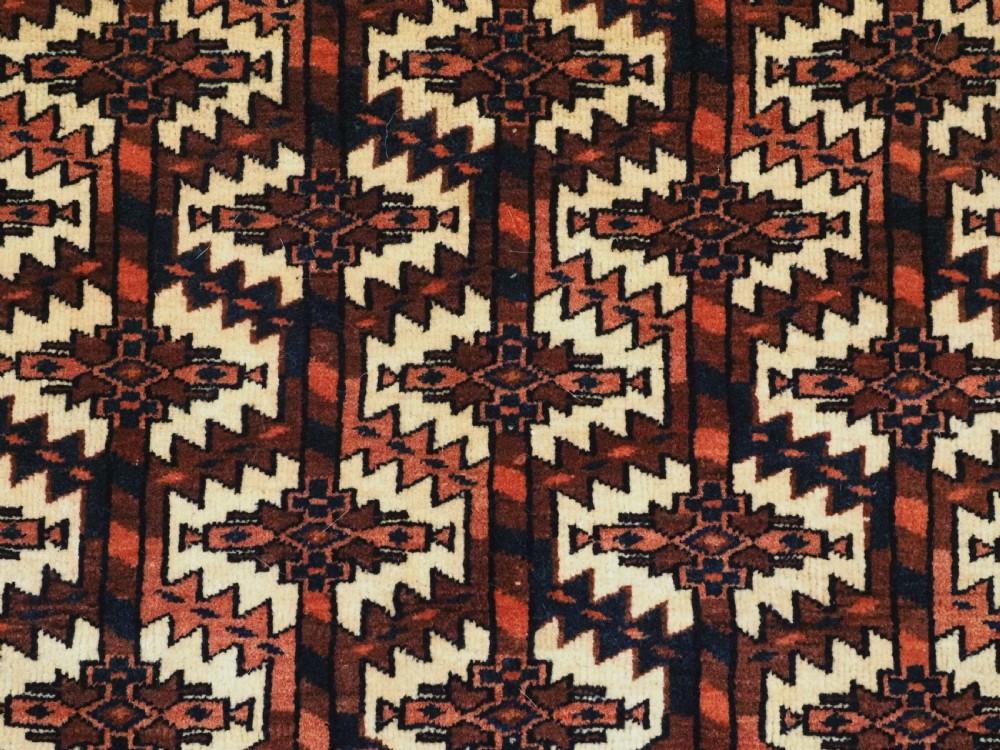 A pentagonal hand-knotted antique Yomut Turkmen Asmalyk wool textile featuring geometric lattice motifs in red, dark brown, and cream with hanging multi-coloured tassels along the lower edge. View VI