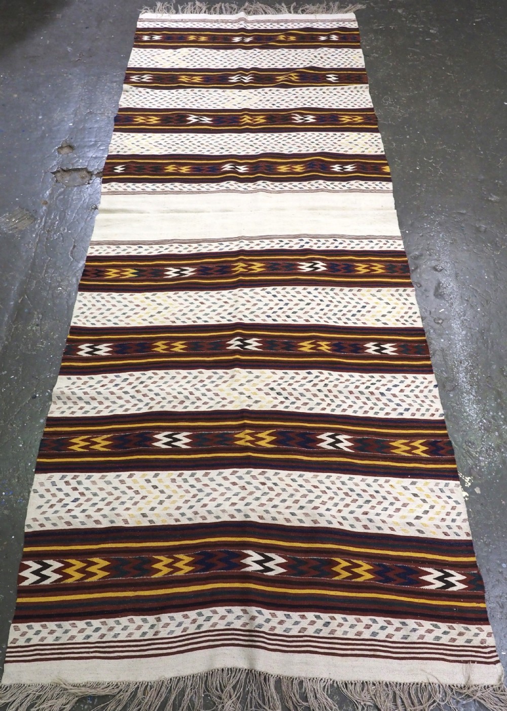 A long handwoven flatweave Central Asian kilim runner displaying cream sections alternating with dark brown, yellow, and red geometric bands with fringe at both ends.