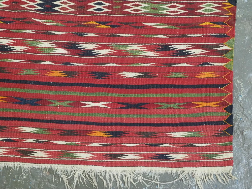 An antique flatweave Turkmen Dali kilim rug featuring horizontal bands of geometric motifs in red, cream, yellow, and dark green shades with fringed ends. View X