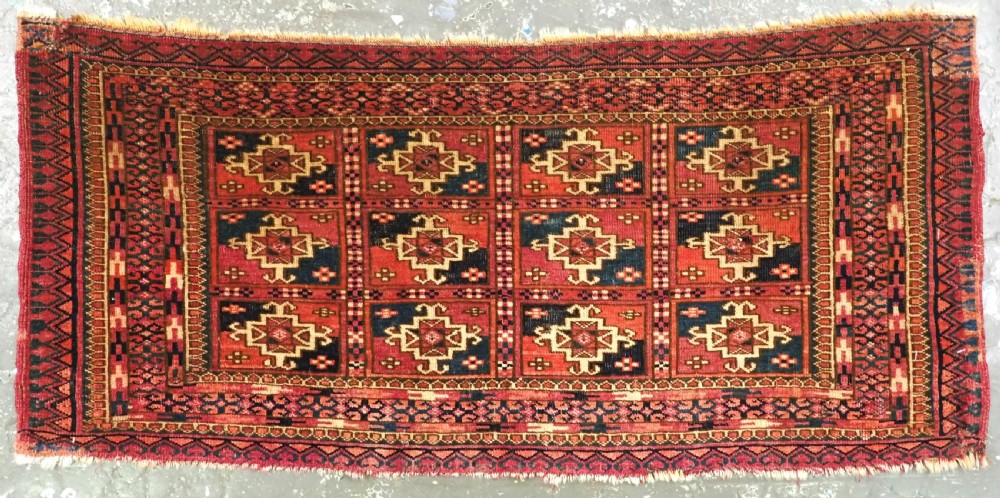 A small rectangular antique Tekke Turkmen mafrash wool panel featuring a repeating grid of geometric aina gul motifs in deep red, dark blue, and cream.