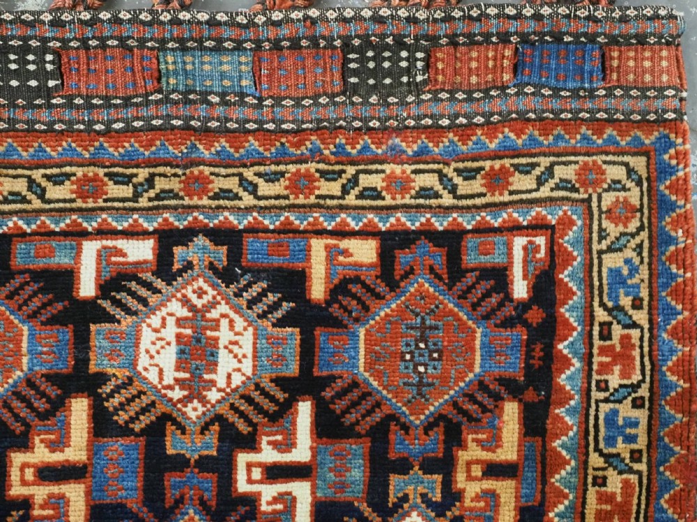 A square handwoven wool Khamseh saddlebag face featuring geometric diamond medallions in terracotta red, cream, and blue set against a dark navy ground with a multi-bordered frame. View IV