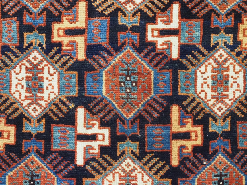 A square handwoven wool Khamseh saddlebag face featuring geometric diamond medallions in terracotta red, cream, and blue set against a dark navy ground with a multi-bordered frame. View VI