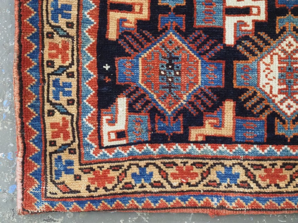 A square handwoven wool Khamseh saddlebag face featuring geometric diamond medallions in terracotta red, cream, and blue set against a dark navy ground with a multi-bordered frame. View VII