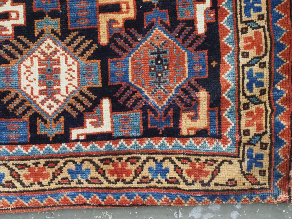 A square handwoven wool Khamseh saddlebag face featuring geometric diamond medallions in terracotta red, cream, and blue set against a dark navy ground with a multi-bordered frame. View VIII