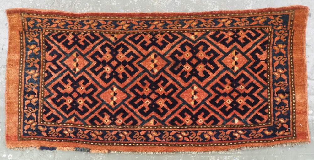 A rectangular antique hand-knotted wool textile featuring geometric navy and orange stepped lattice motifs on a rust-red field framed by a patterned border.