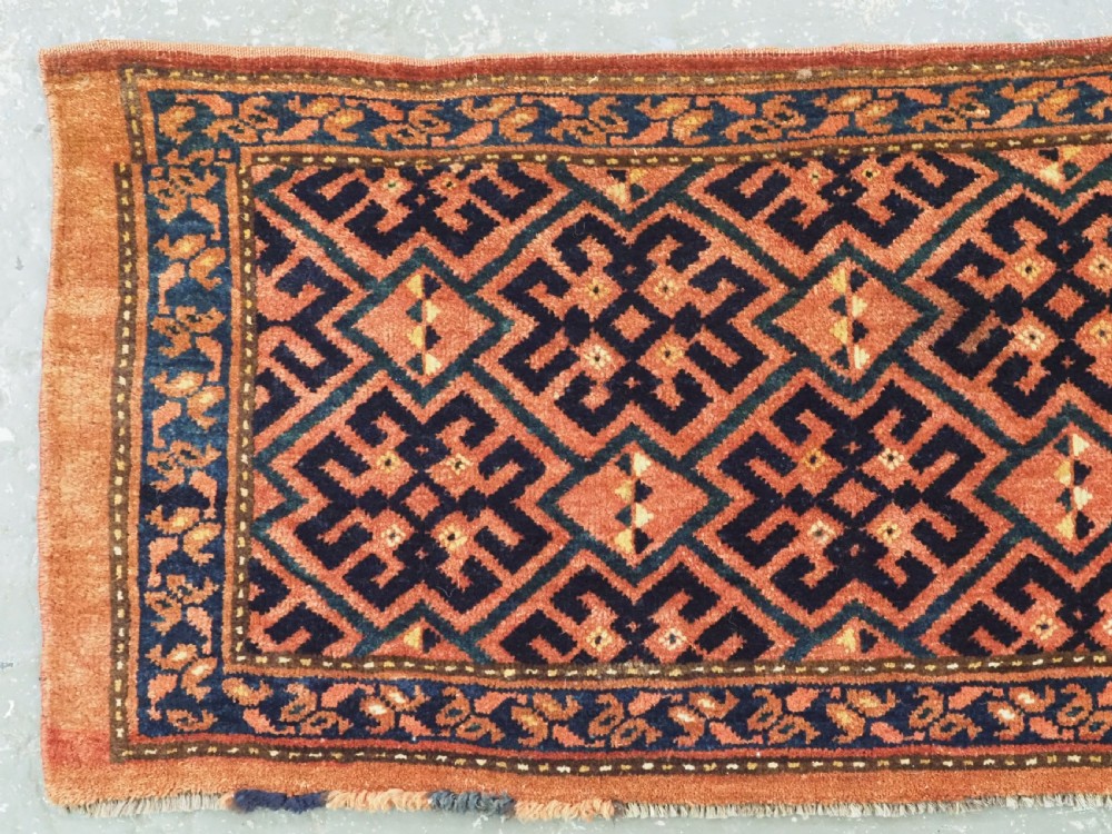 A rectangular antique hand-knotted wool textile featuring geometric navy and orange stepped lattice motifs on a rust-red field framed by a patterned border. View II