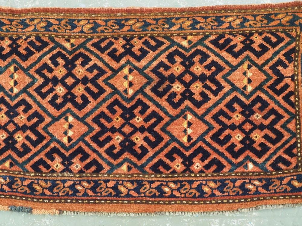 A rectangular antique hand-knotted wool textile featuring geometric navy and orange stepped lattice motifs on a rust-red field framed by a patterned border. View III