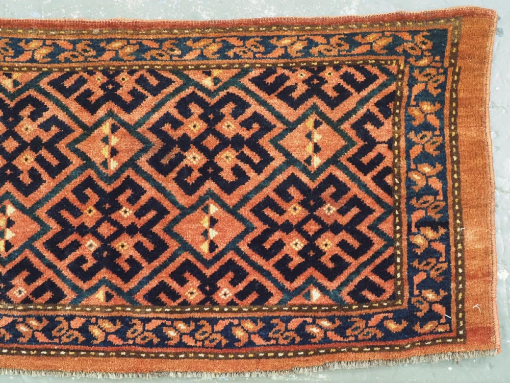 A rectangular antique hand-knotted wool textile featuring geometric navy and orange stepped lattice motifs on a rust-red field framed by a patterned border. View IV