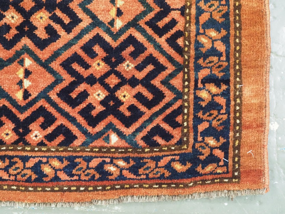 A rectangular antique hand-knotted wool textile featuring geometric navy and orange stepped lattice motifs on a rust-red field framed by a patterned border. View V