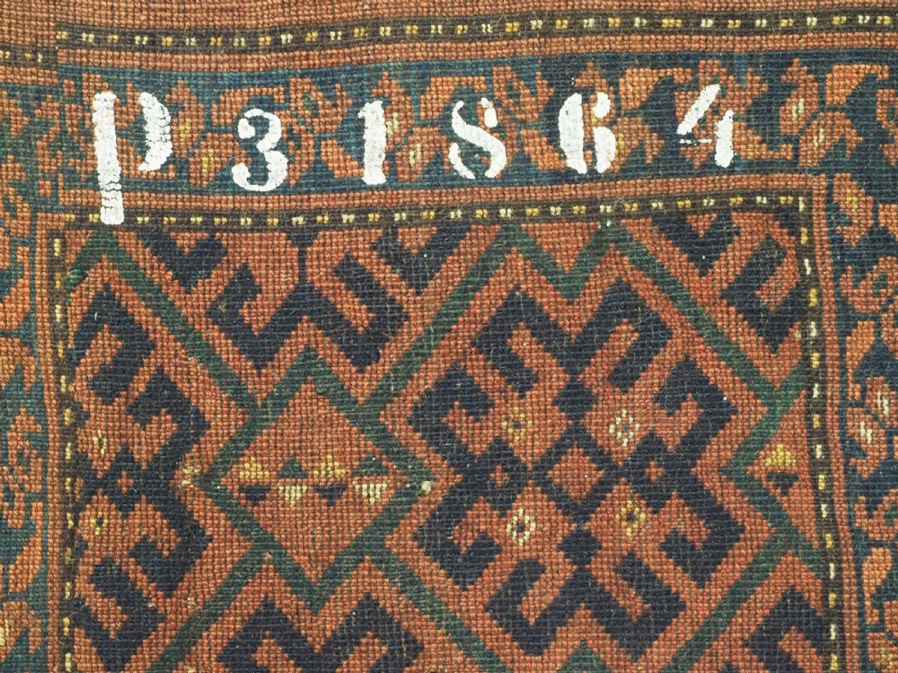 A rectangular antique hand-knotted wool textile featuring geometric navy and orange stepped lattice motifs on a rust-red field framed by a patterned border. View VI