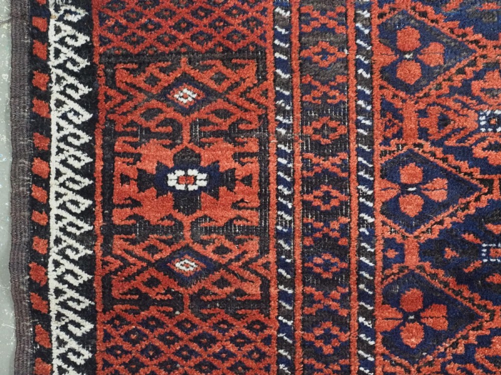 An antique rectangular Khorasan Baluch hand-knotted wool rug featuring repeating central dark blue geometric diamond motifs framed by multiple terracotta red pattern borders. View VII