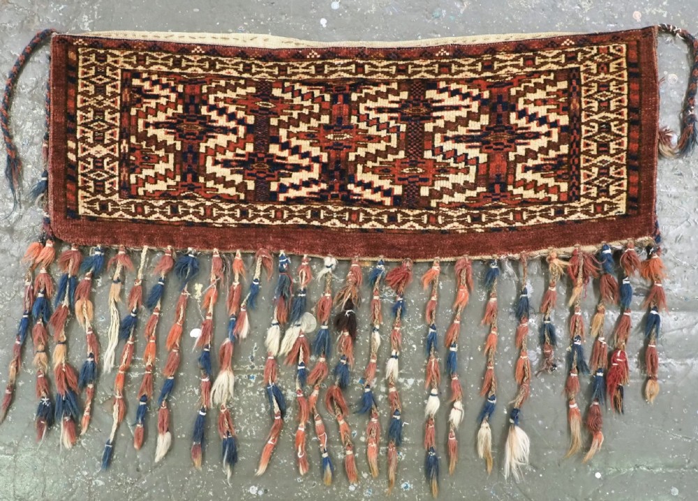 An antique rectangular Yomut Turkmen wool textile featuring intricate geometric lattice patterns in terracotta red, navy blue, and ivory, finished with a fringe of multi-coloured tassels along the bottom edge.