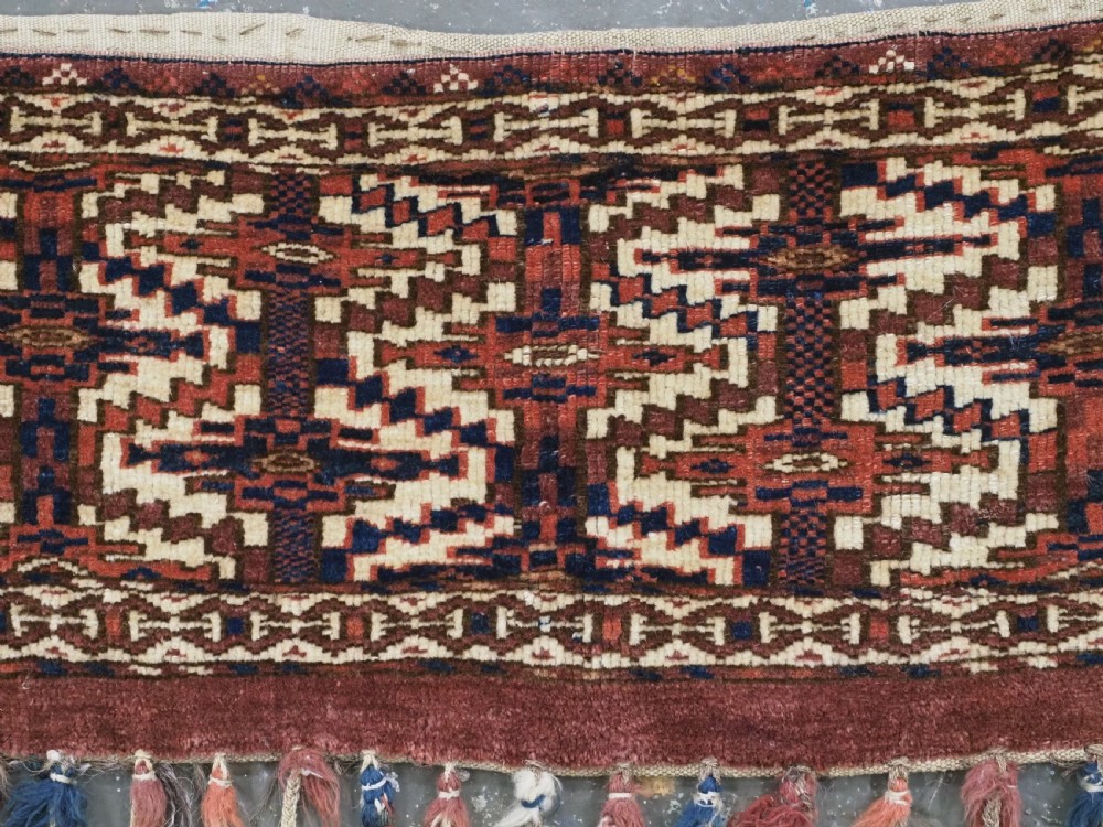 An antique rectangular Yomut Turkmen wool textile featuring intricate geometric lattice patterns in terracotta red, navy blue, and ivory, finished with a fringe of multi-coloured tassels along the bottom edge. View III