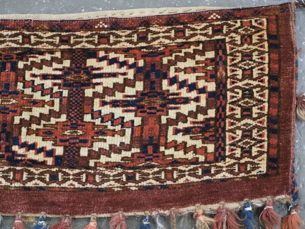 An antique rectangular Yomut Turkmen wool textile featuring intricate geometric lattice patterns in terracotta red, navy blue, and ivory, finished with a fringe of multi-coloured tassels along the bottom edge. View IV
