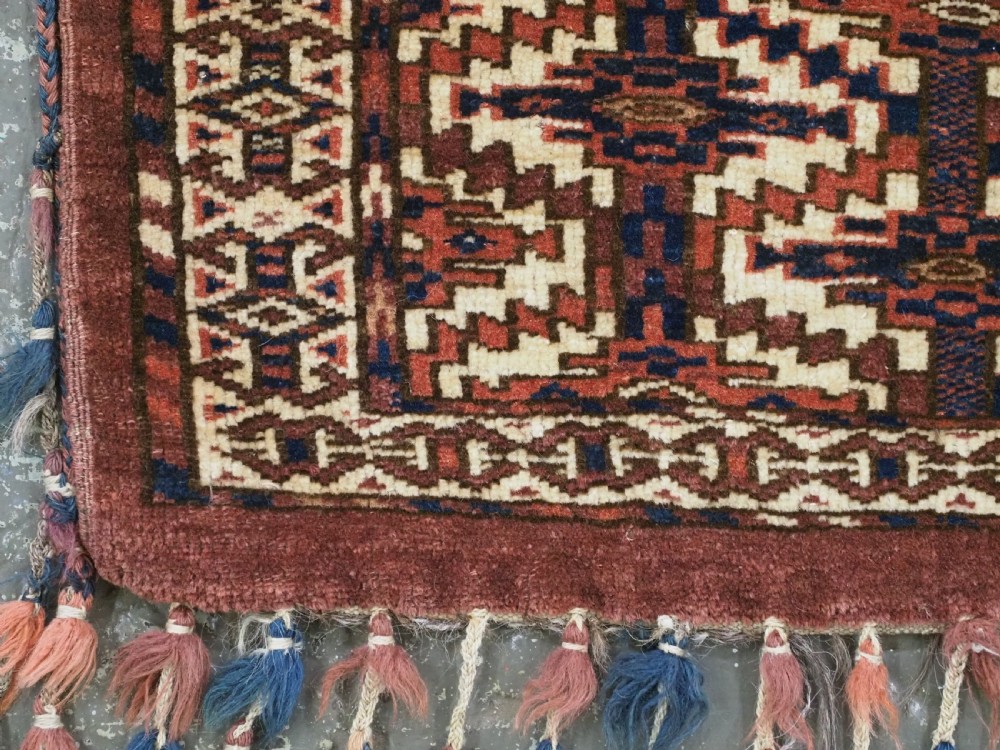 An antique rectangular Yomut Turkmen wool textile featuring intricate geometric lattice patterns in terracotta red, navy blue, and ivory, finished with a fringe of multi-coloured tassels along the bottom edge. View V