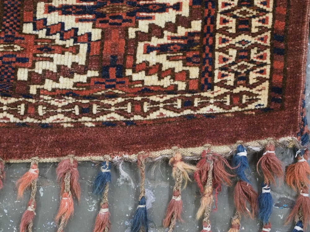 An antique rectangular Yomut Turkmen wool textile featuring intricate geometric lattice patterns in terracotta red, navy blue, and ivory, finished with a fringe of multi-coloured tassels along the bottom edge. View VI