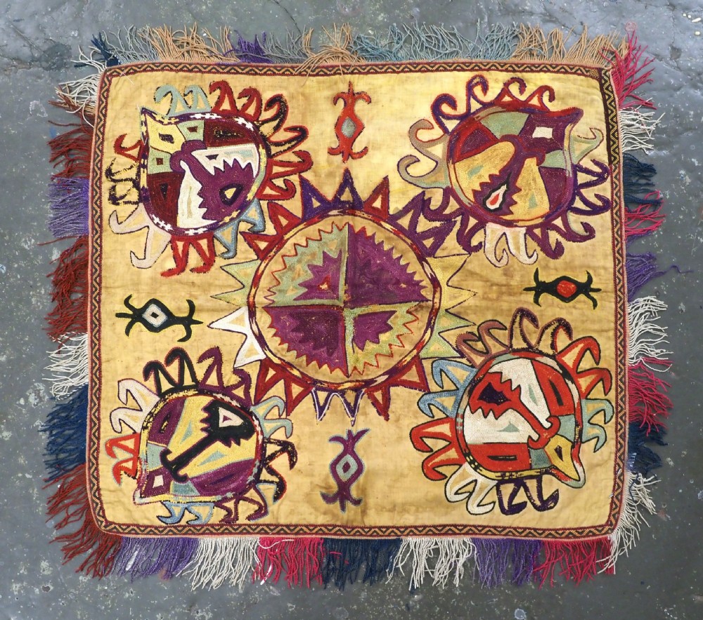 A square antique Uzbek Lakai embroidered textile featuring central circular and solar motifs in purple, red, and turquoise on a yellow fabric ground, finished with a multi-coloured fringed border.
