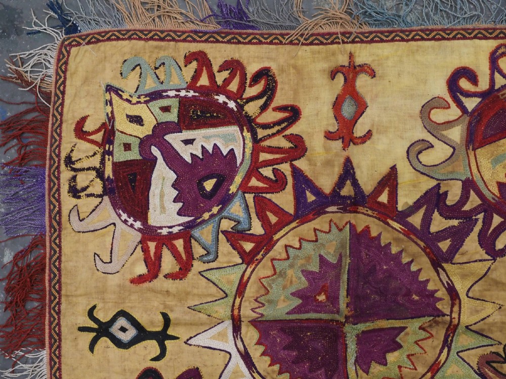 A square antique Uzbek Lakai embroidered textile featuring central circular and solar motifs in purple, red, and turquoise on a yellow fabric ground, finished with a multi-coloured fringed border. View II