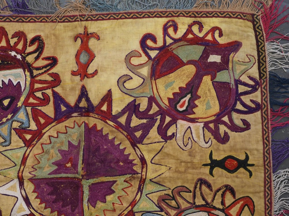A square antique Uzbek Lakai embroidered textile featuring central circular and solar motifs in purple, red, and turquoise on a yellow fabric ground, finished with a multi-coloured fringed border. View III